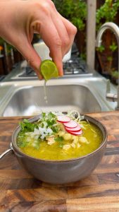 Hand squeezing fresh lime juice over pozole verde with toppings.