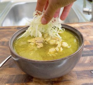 Hand placing shredded cabbage on top of pozole verde.