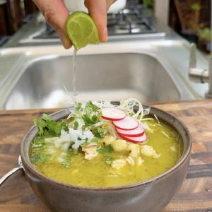 Hand squeezing fresh lime into a bowl of Chicken Pozole Verde topped with cabbage, radish, and cilantro.