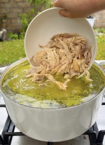 Shredded cooked chicken being poured into the pot of green pozole.