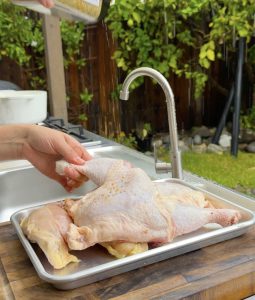 Hand sprinkling seasoning over raw chicken pieces on a tray.