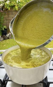 Blended green salsa being poured into the pot of broth and hominy.